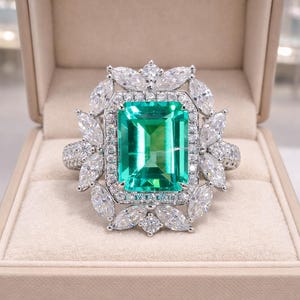 Puede incluir: Un gran anillo con una piedra verde talla esmeralda, rodeada de piedras blancas y una banda con diamantes. El anillo, engastado en plata u oro blanco, se presenta en un joyero beige.
