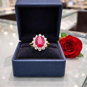May include: A gold ring with a large oval ruby center stone, surrounded by a halo of smaller diamonds. The ring is displayed in a blue velvet box, with a red rose in the background.