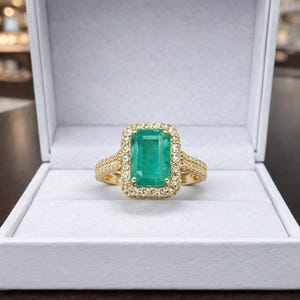 May include: A gold ring featuring a large emerald-cut green gemstone, encircled by smaller diamonds. The ring is set in a gold band and displayed in a white jewellery box. The design of the ring is classic and elegant.