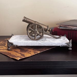 Può includere: Un cannone in miniatura color bronzo è posto su un vassoio bianco effetto marmo con piedini in metallo ornati. Il cannone ha ruote dettagliate e una canna, suggerendo un tema storico o decorativo. Il vassoio è su una superficie scura.