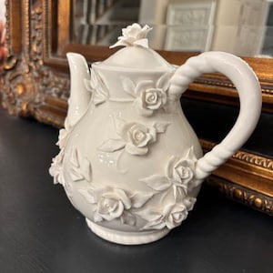 May include: Cream-colored ceramic teapot with a floral rose design. The teapot features a curved handle and a spout, with roses and leaves sculpted on the body and lid. The teapot is a decorative item.