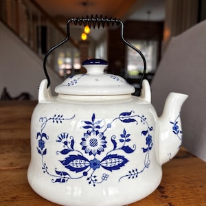 May include: White ceramic teapot with a blue floral design. The teapot has a black metal handle and a matching lid. The floral pattern includes flowers and leaves. The teapot is sitting on a wooden surface.