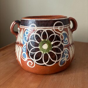 May include: A terracotta pot with two side handles, featuring a dark brown rim and a hand-painted floral design in white, blue, and green. The body of the vessel is a warm reddish-brown clay, decorated with symmetrical white line patterns.