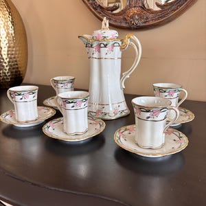 May include: A white porcelain tea set with a coffee pot, six cups, and saucers. The set features pink floral designs, gold trim, and a decorative handle. The cups and saucers are small, and the coffee pot is tall.