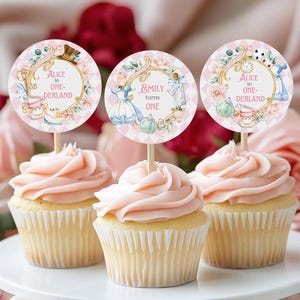 Könnte beinhalten: Drei Cupcakes mit rosa Zuckerguss und dekorativen Toppern. Die Topper zeigen Alice im Wunderland-Motive mit dem Text "Alice in One-derland" und "Emily turns one". Der Hintergrund ist ein zartes Rosa.