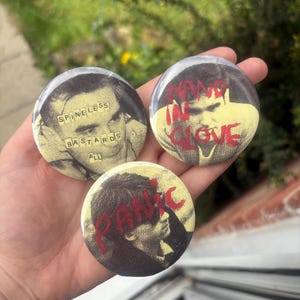 May include: Three round pinback buttons with black and white images and text. One button reads "SPINELESS BASTARDS ALL", another says "HAND IN GLOVE", and the third says "PANIC". The buttons are held in a hand.