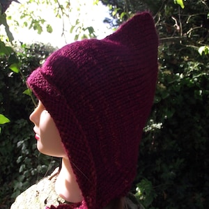 Può includere: Un primo piano di un cappello con cappuccio in maglia color borgogna con punta a punta. Il cappello è indossato da una persona con i capelli lunghi e castani. Il cappello è fatto di un filato morbido e testurizzato.