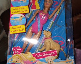 Könnte beinhalten: Ein Barbie Swim Champion Puppenset in Originalverpackung mit einer Puppe im pinkfarbenen Badeanzug mit der Nummer 59, einer Golden Retriever Welpenfigur, einem pinkfarbenen Pokal und Texten wie Team Barbie, Wind & watch doll really swim!, Pup really swims too!.