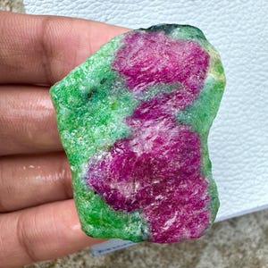 May include: A rough-cut gemstone featuring vibrant magenta ruby inclusions within a matrix of green zoisite. The natural stone displays a mix of textures and colors, showcasing the raw beauty of the mineral. The stone is held in a hand.
