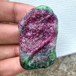 May include: A raw ruby gemstone, showcasing a deep magenta interior and a textured surface. The edges have green and grey tones, giving a natural look. The gemstone is held in a hand.