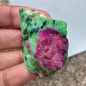 Peut inclure: Une pierre brute de rubis dans du zoïsite. La pierre présente un mélange de couleurs vives, avec une section de rubis magenta foncé sur une matrice de zoïsite verte. Des mouchetures noires sont dispersées dans les zones vertes.