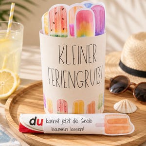May include: A cylindrical paper container featuring the text KLEINER FERIENGRUB and watercolor popsicle illustrations. A separate paper strip displays the text du kannst jetzt die Seele baumeln lassen! alongside a single orange popsicle graphic.