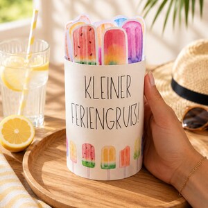 May include: A cylindrical white paper container featuring the black text "KLEINER FERIENGRUB!" and colorful watercolor illustrations of various popsicle shapes. The container is filled with several die-cut popsicle-shaped paper cards.
