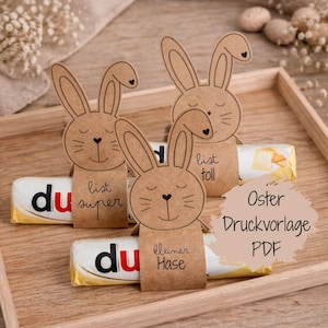 Puede incluir: Tres decoraciones de conejos de papel marrón con caras dibujadas, sujetas a barras de chocolate. Las barras de chocolate tienen envoltorios blancos y amarillos con la palabra "du" en rojo. El texto "Oster Druckvorlage PDF" está en un círculo marrón claro.