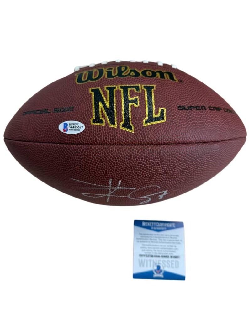 May include: A brown Wilson NFL football with white laces and gold lettering. The football has a signature and a Beckett authentication sticker. A Beckett Certificate of Authenticity is below the football.