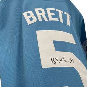 May include: Light blue jersey with the name "BRETT" and the number "5" in white. The jersey has a signature below the number. The collar is white and blue.