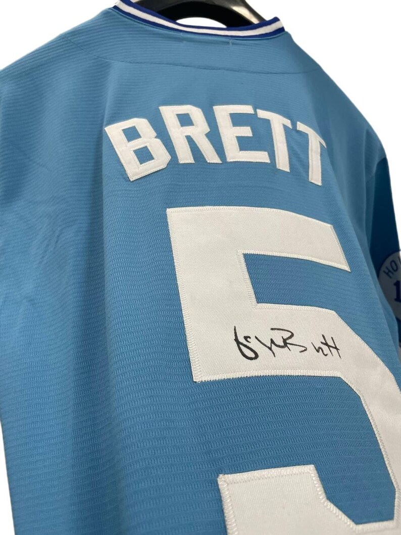 May include: Light blue jersey with the name "BRETT" and the number "5" in white. The jersey has a signature below the number. The collar is white and blue.