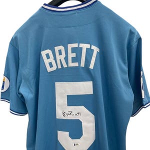 May include: Light blue baseball jersey with the name "BRETT" and the number "5" in white on the back. The jersey has a signature below the number. The collar and sleeves have white and blue trim.