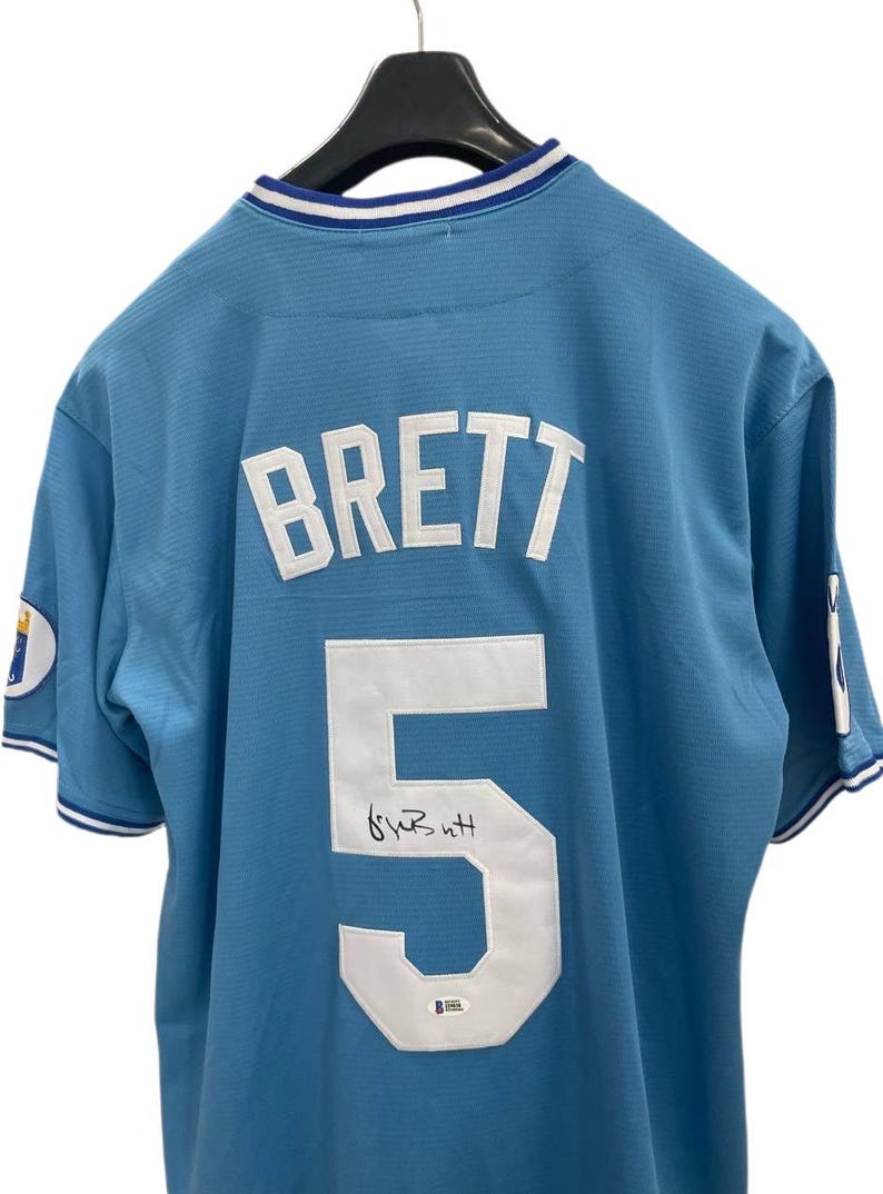 May include: Light blue baseball jersey with the name "BRETT" and the number "5" in white on the back. The jersey has a signature below the number. The collar and sleeves have white and blue trim.