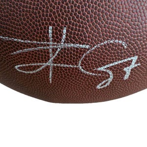 May include: Close-up of a brown football with white stitching and a silver signature. The football has a textured surface and is partially visible against a white background. The signature is in cursive.
