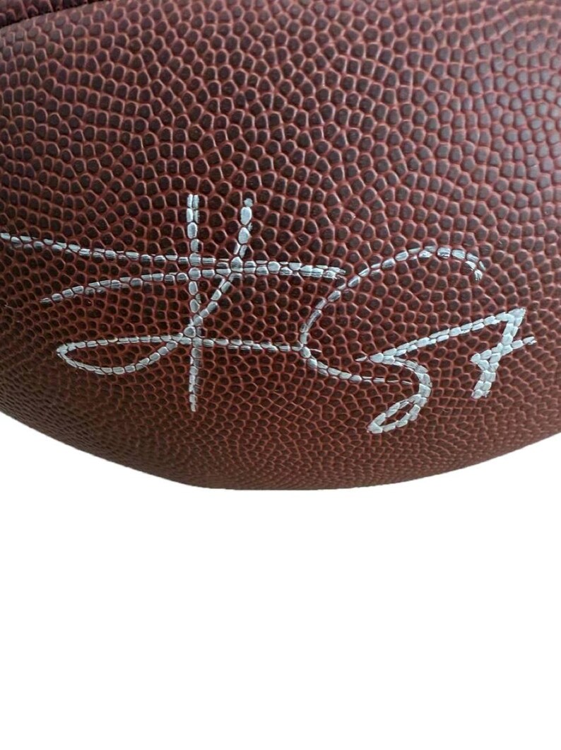 May include: Close-up of a brown football with white stitching and a silver signature. The football has a textured surface and is partially visible against a white background. The signature is in cursive.