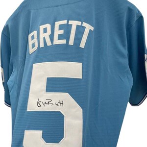 May include: Light blue baseball jersey with the name "BRETT" and the number "5" in white. The jersey has a signature below the number and a Beckett Authentication sticker.