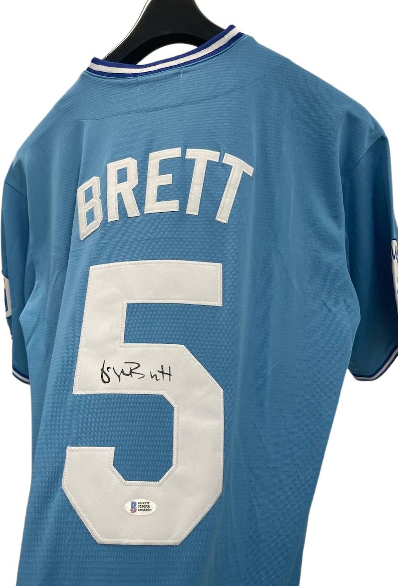 May include: Light blue baseball jersey with the name "BRETT" and the number "5" in white. The jersey has a signature below the number and a Beckett Authentication sticker.