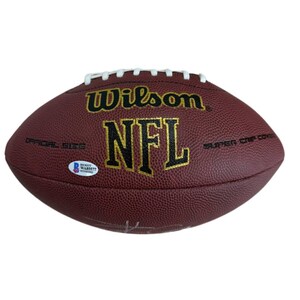 May include: A maroon Wilson NFL football with white laces. The football has the NFL logo in gold lettering. The words "Wilson" and "Super Grip Cover" are also visible. A Beckett Authentication Services sticker is on the football.