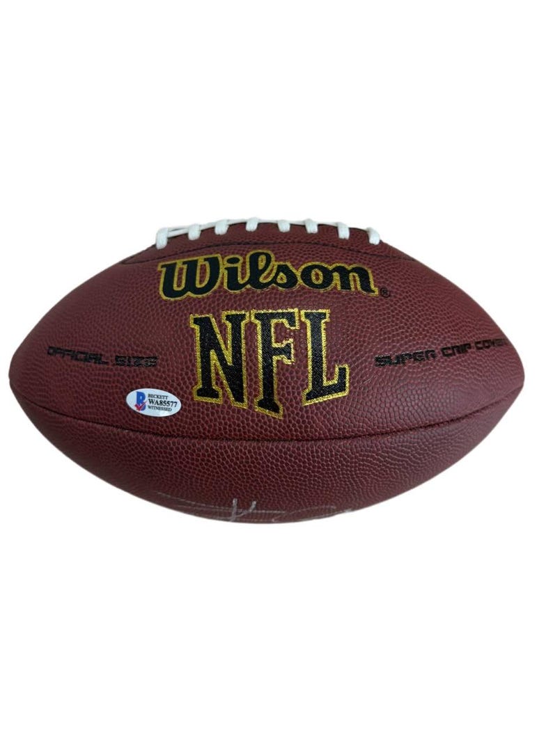 May include: A maroon Wilson NFL football with white laces. The football has the NFL logo in gold lettering. The words "Wilson" and "Super Grip Cover" are also visible. A Beckett Authentication Services sticker is on the football.