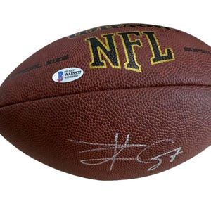 May include: A brown NFL football with the text "NFL" in gold lettering. The football has a textured surface and a white sticker with text. A silver signature is visible on the football.