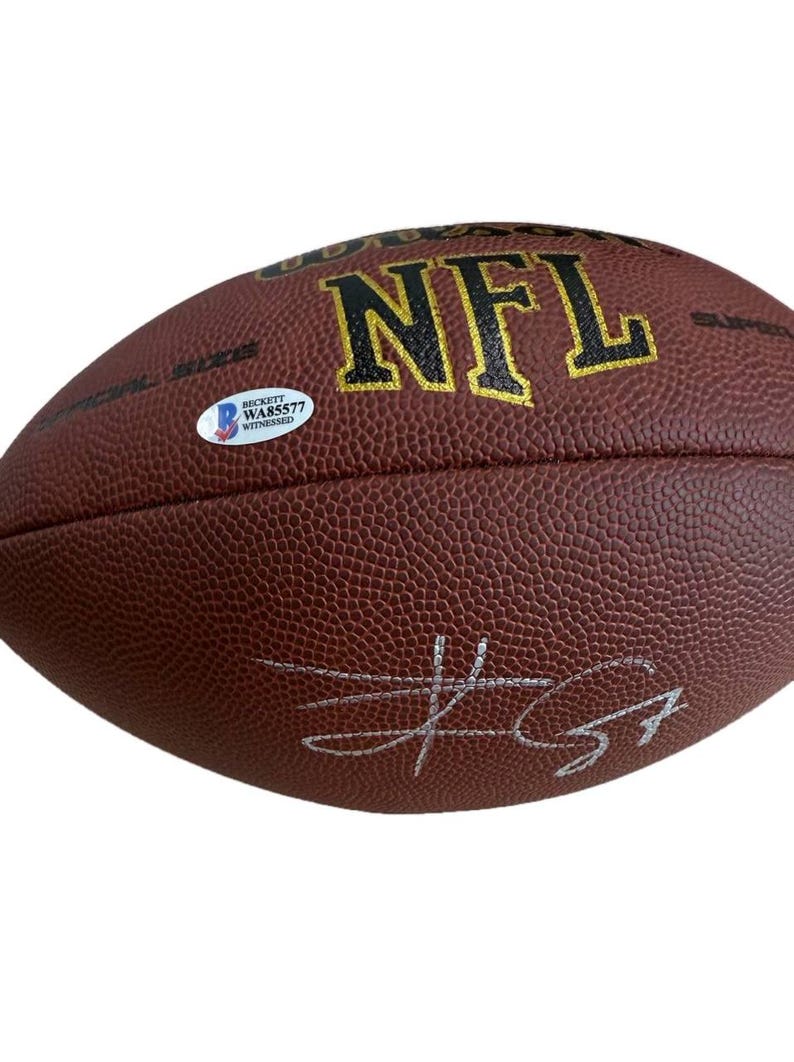 May include: A brown NFL football with the text "NFL" in gold lettering. The football has a textured surface and a white sticker with text. A silver signature is visible on the football.