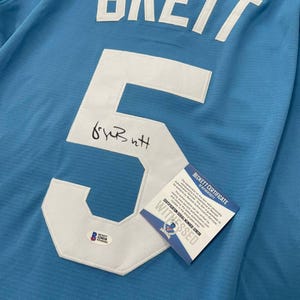 May include: Light blue jersey with the name "BRETT" in white letters across the top. A large white number "5" is sewn on the front, with a signature below. A Beckett Certificate of Authenticity is attached.
