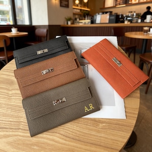 May include: An assortment of leather wallets in various colors: black, brown, and orange. Each wallet features a rectangular shape with a flap closure and a silver-colored clasp. One wallet has the initials "A.R." in gold.