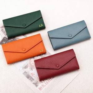 May include: Four leather wallets in shades of green, orange, blue, and burgundy. Each wallet has a flap closure with a button and the initials "A.R." in gold. The wallets are arranged on a newspaper.