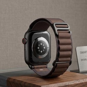 May include: A black smartwatch with a brown fabric band is displayed on a wooden stand. The watch face is dark, and the band has a textured appearance. The watch is a modern accessory.