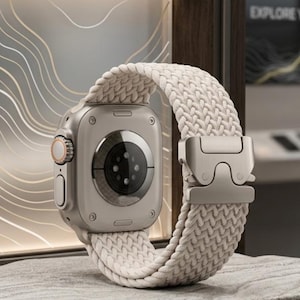 Op de afbeelding: Een zilveren smartwatch met een crèmekleurige geweven band. Het horloge heeft een digitaal display en een zijknop. De band heeft een metalen sluiting. Het horloge wordt getoond op een gestructureerd, grijs oppervlak.