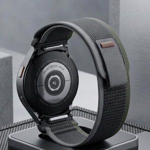 May include: A black Galaxy Watch with a gray and olive green fabric band rests on a black charging stand. The watch face is visible, and the band is looped around the stand. The watch is a modern accessory.