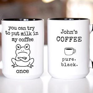 May include: Two white ceramic mugs with black handles and interiors. The left mug features a drawing of a grumpy frog with the text: you can try to put milk in my coffee once. The right mug displays the text: John's COFFEE, a small cup icon, and pure. black.