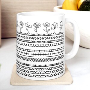 May include: A white ceramic mug featuring a black line art design. The pattern consists of a top row of simple flower illustrations followed by several horizontal bands of geometric shapes, including triangles, scallops, squares, and zigzags.