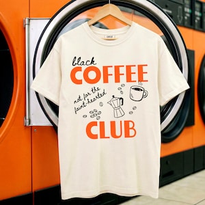 以下が含まれることがあります： 黒とオレンジ色の「black COFFEE」の文字とコーヒー関連のイラストが描かれたクリーム色のTシャツ。「not for the faint-hearted CLUB」の文字も表示されています。木製のハンガーにTシャツが掛けられています。