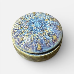 May include: A round, ceramic trinket box with a lid. The lid features a blue and gold speckled pattern. The box has a brass-colored band around the base and lid. The box is a decorative item.
