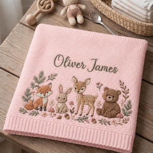 May include: A soft pink knitted baby blanket featuring the embroidered name Oliver James in dark thread. The blanket displays a detailed woodland scene with a fox, a rabbit, a fawn, and a bear surrounded by small flowers, mushrooms, and pine branches.