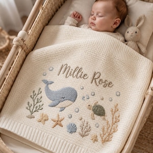May include: A cream-coloured knit baby blanket featuring embroidered ocean-themed designs including a blue whale, a green sea turtle, starfish, seashells, and seaweed. The name Millie Rose is embroidered in brown script above the whale.