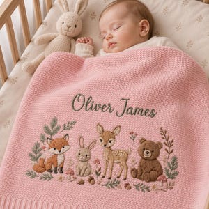 May include: A soft pink knitted baby blanket featuring the embroidered name Oliver James in dark green script. Below the name is a detailed woodland scene with a fox, a rabbit, a fawn, and a bear surrounded by small flowers, leaves, and mushrooms.