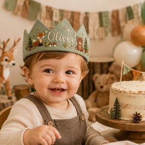 May include: A sage green felt crown featuring embroidered forest elements including a fox, a bear, pine trees, mushrooms, and small stars. The name Oliver is embroidered in white script in the centre of the crown, which has a pointed top edge.