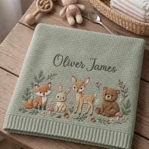 May include: A sage green knitted baby blanket featuring the embroidered name Oliver James in dark green script. The blanket displays a detailed woodland scene with an embroidered fox, rabbit, fawn, and bear surrounded by small flowers and mushrooms.