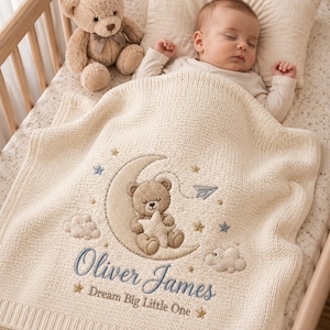 Può includere: Una coperta per neonati in maglia color crema con un ricamo raffigurante un orsetto seduto su una luna crescente che tiene in mano una stella. La coperta presenta il testo ricamato "Oliver James" e "Dream Big Little One" in blu e oro.