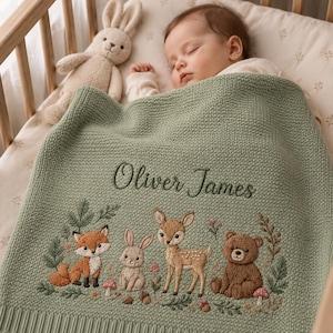 May include: A sage green knitted baby blanket featuring the embroidered name Oliver James in dark green script. The blanket displays a detailed woodland scene with a fox, a rabbit, a fawn, and a bear surrounded by small flowers and mushrooms.