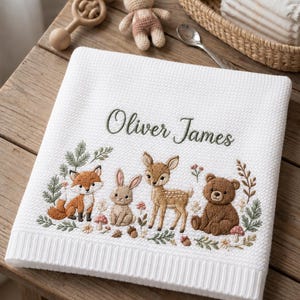 May include: A white textured baby blanket featuring the embroidered name Oliver James in green script. Below the name is a detailed woodland scene with an embroidered fox, rabbit, fawn, and bear surrounded by small flowers, mushrooms, and greenery.