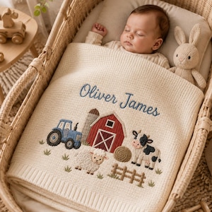 May include: A cream-colored knit baby blanket featuring an embroidered farm scene with a blue tractor, a red barn, a sheep, and a cow. The name Oliver James is embroidered in blue script above the farm imagery. The blanket has a ribbed border.